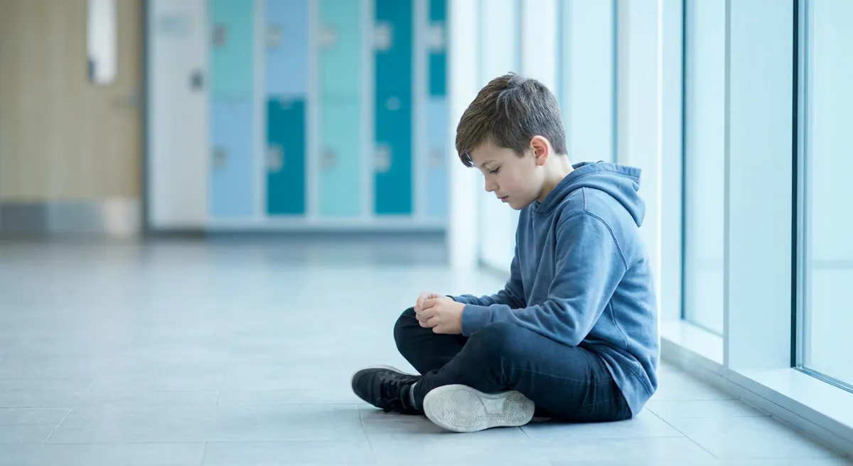 Niño sentado solo en el suelo de un pasillo escolar con actitud introspectiva y triste