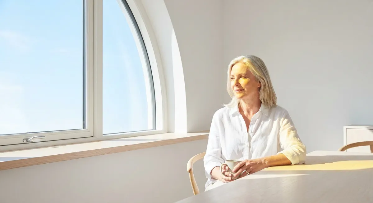 Mujer madura en calma frente a una ventana reflejando equilibrio emocional durante la menopausia